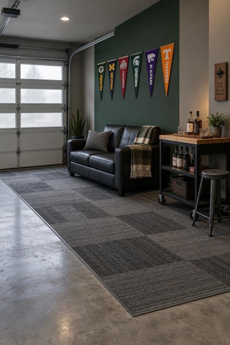 11 Cozy Sports Man Cave Ideas for Game-Day Comfort 7 A garage bay seating zone with gray carpet tile over a concrete slab, two painted wall panels with a hunter green block, a pennant run, a black leather loveseat, and a rolling bar cart in cool daylight.