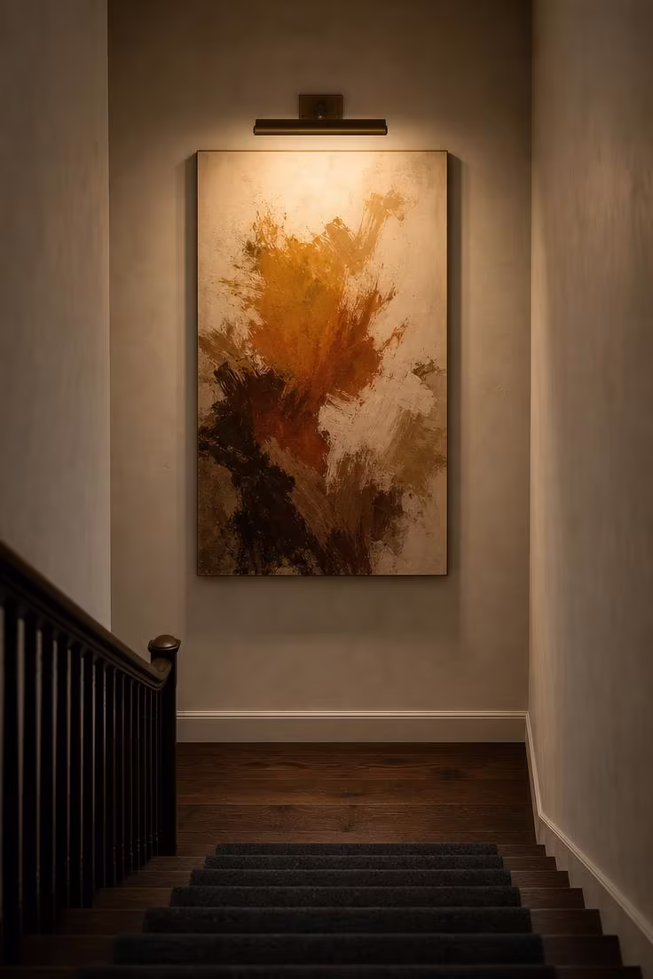 Oversized abstract canvas in earth tones on a stair landing wall lit by an aged brass picture light