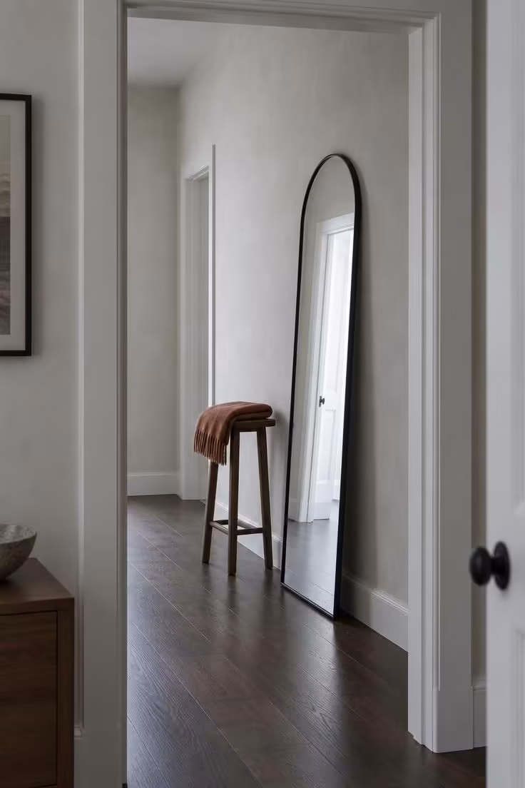 17 Cozy Stairs and Hallway Ideas You'll Want to Steal 13 A tall narrow arched mirror leaning against a hallway wall beside a slim wooden stool with a folded throw.