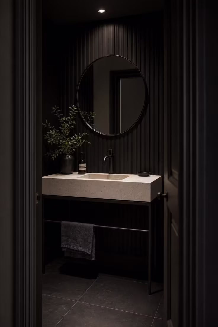 Doorway view into a dark statement powder room with black fluted wood paneling, a honed limestone console vanity on iron legs, and moody low lighting.
