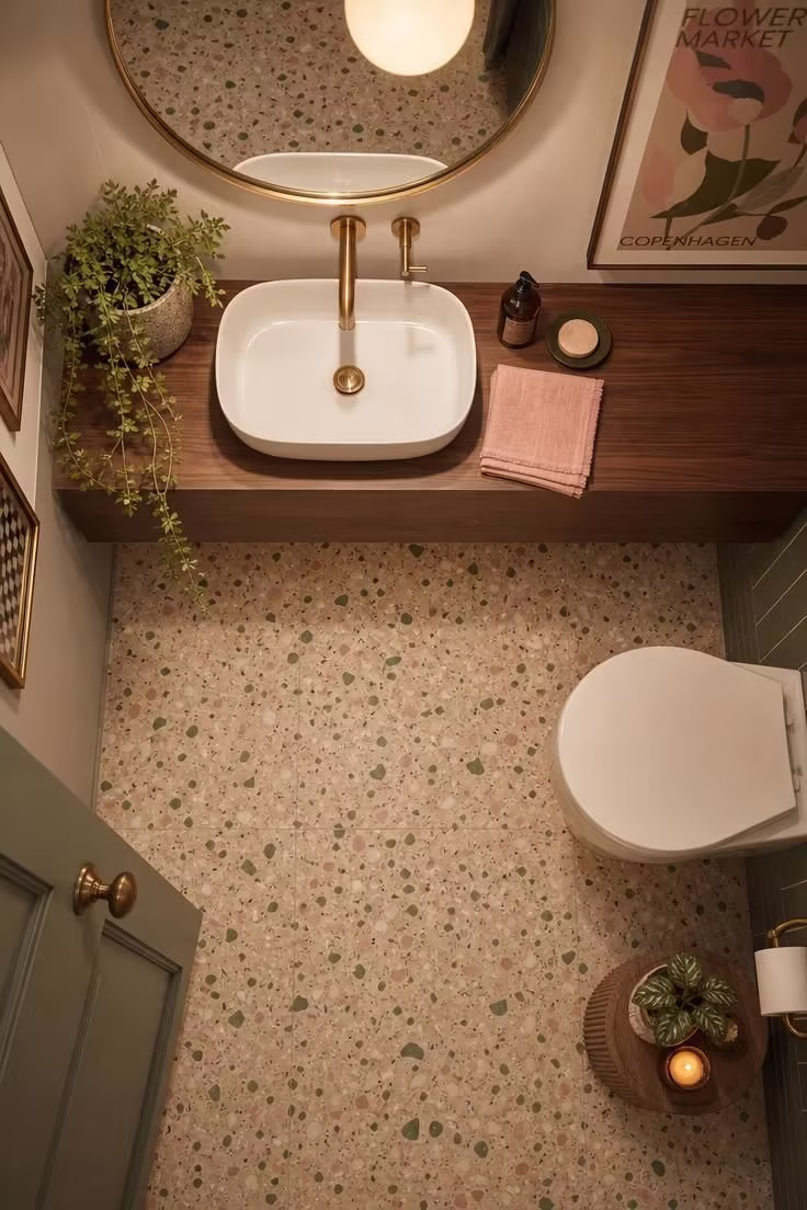 Overhead view of a terrazzo floor with sage and blush chips, a walnut floating vanity, a shallow white basin, and a round brass-rimmed mirror in a statement powder room.