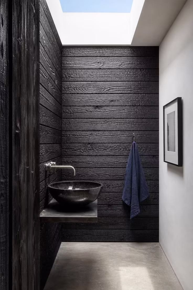 Eye-level view of a shou sugi ban charred cedar accent wall with a black granite vessel basin on a raw steel shelf, lit by bright skylight in a statement powder room.
