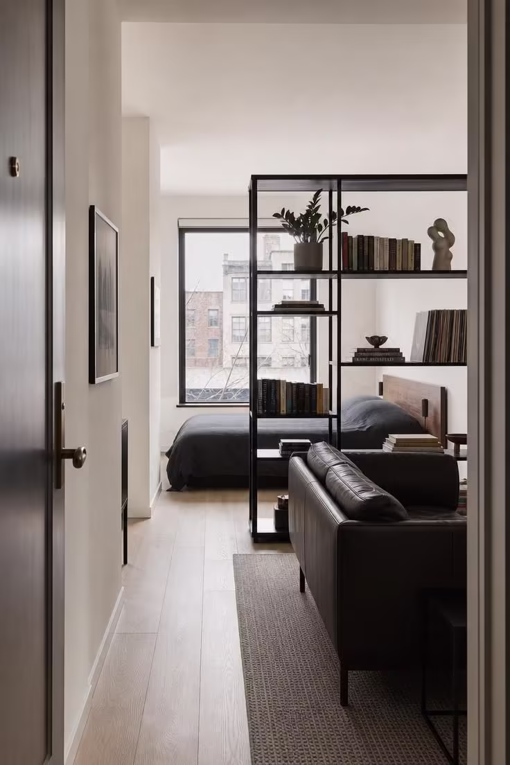 Matte black bookshelf turned sideways as a room divider between living and sleeping zones in a cool-lit studio apartment