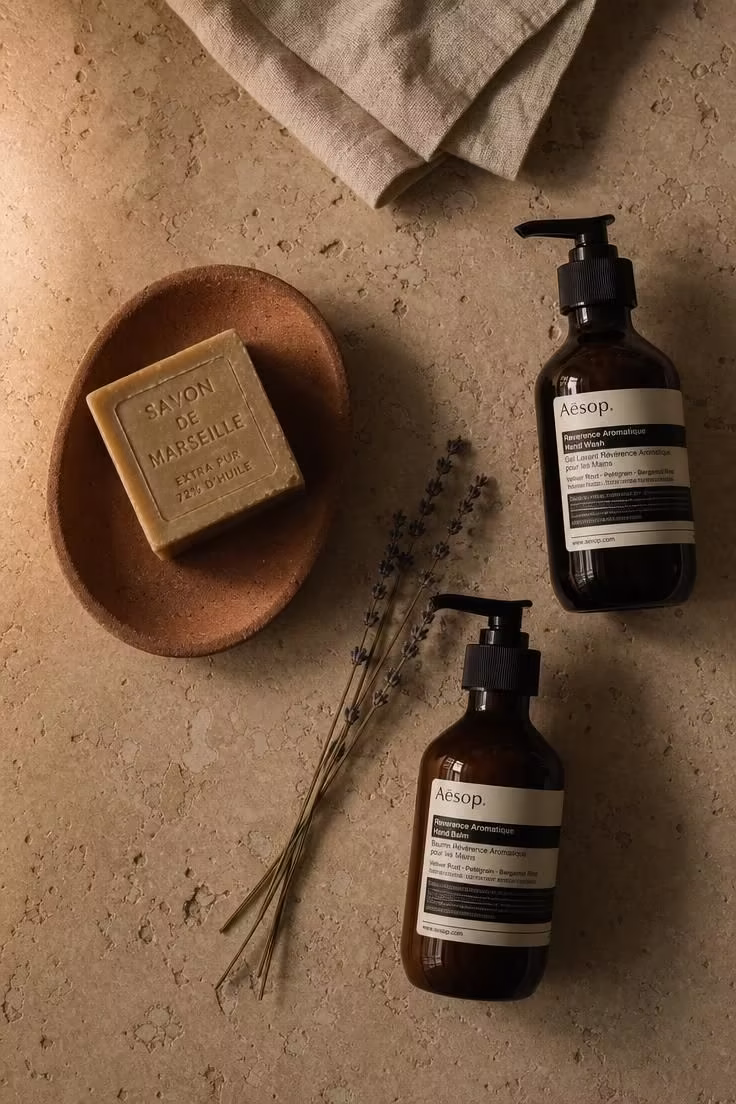 Overhead view of terracotta soap dish, amber glass bottles, and dried lavender on a travertine countertop in moody low light