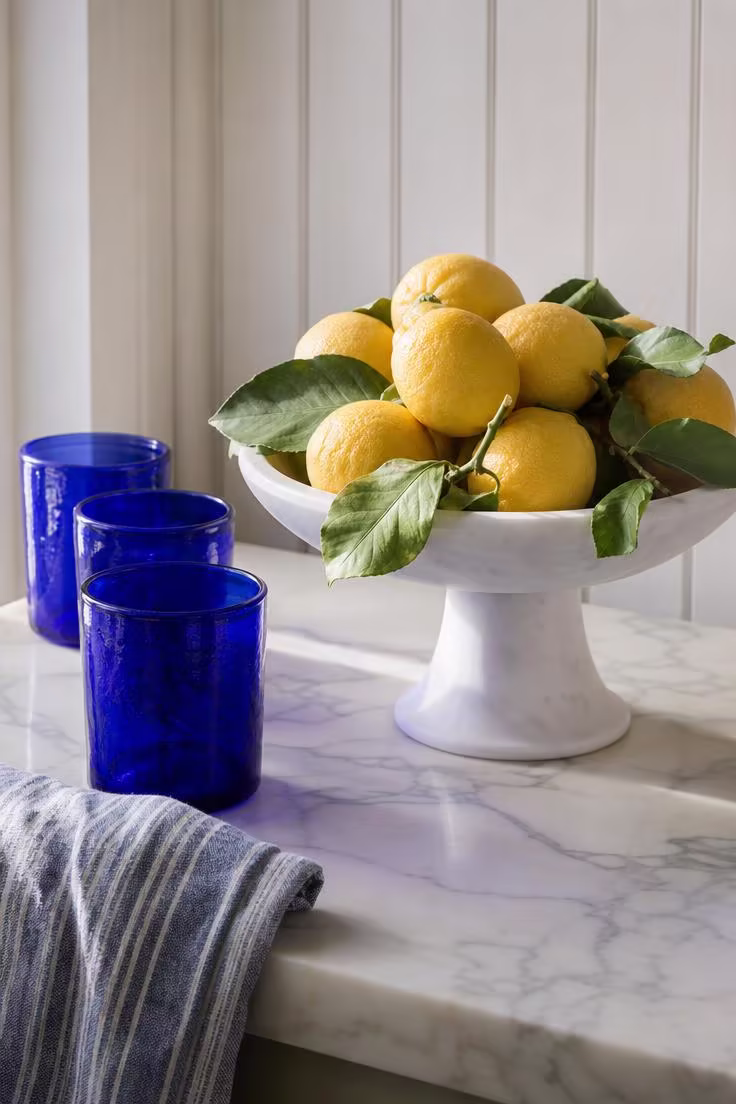 15 Bright Summer Kitchen Ideas to Refresh Your Space 1 Meyer lemons piled on a white marble pedestal next to cobalt blue glassware on a honed Calacatta counter.