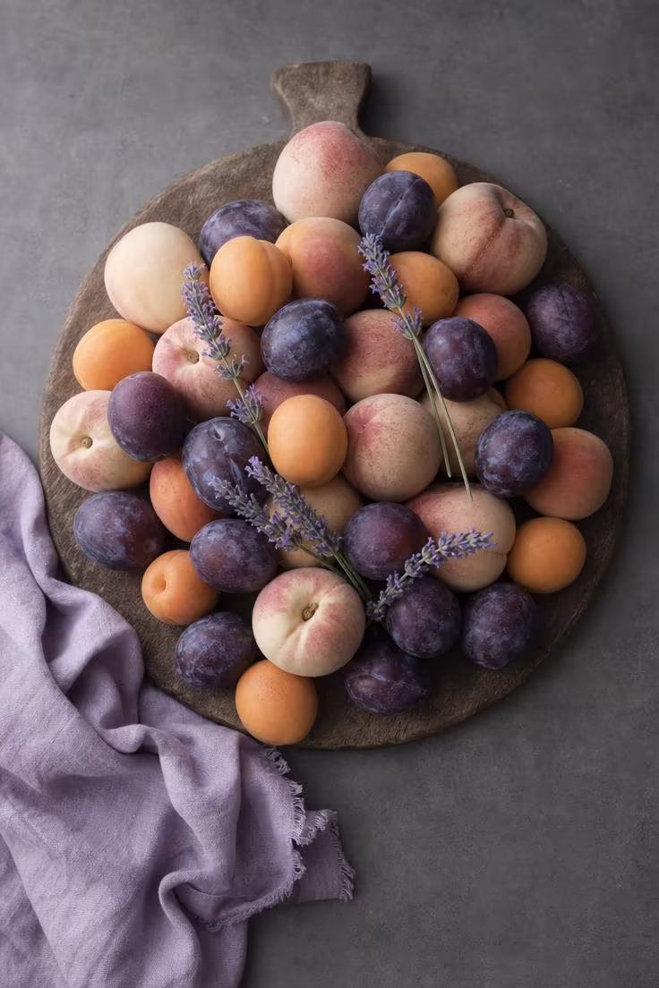 15 Bright Summer Kitchen Ideas to Refresh Your Space 7 A footed wooden bread board piled with white peaches, plums, apricots, and lavender sprigs on a slate gray counter.