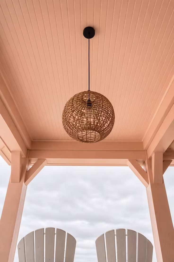 Pale peach painted beadboard porch ceiling with a large rattan globe pendant light hanging from a black cord shot from below on an overcast day