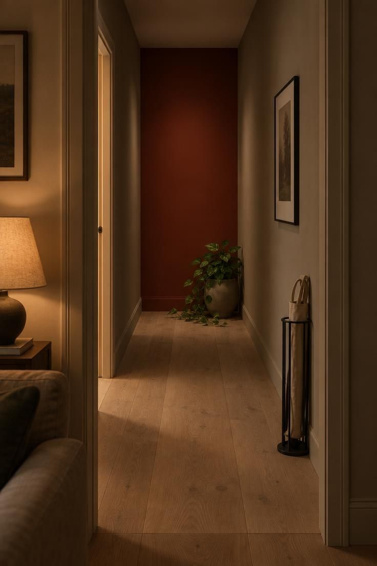 11 Warm Two Tone Hallway Ideas for Small Entryways 4 Short hallway seen from a living room doorway, with a deep clay-red end wall and warm greige side walls under low evening lamplight and pale oak flooring.
