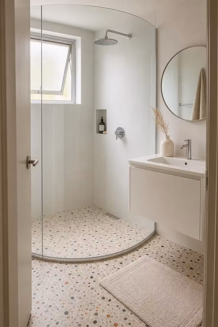 13 Cozy Very Small Bathroom Ideas With Shower Worth Trying 9 A curved glass shower enclosure in the corner of a very small bathroom with a terrazzo-look porcelain base, white tile walls, and soft diffused light from a frosted window.