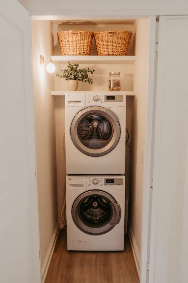 A stacked washer and dryer set inside a bright, narrow laundry alcove with floating shelves above
