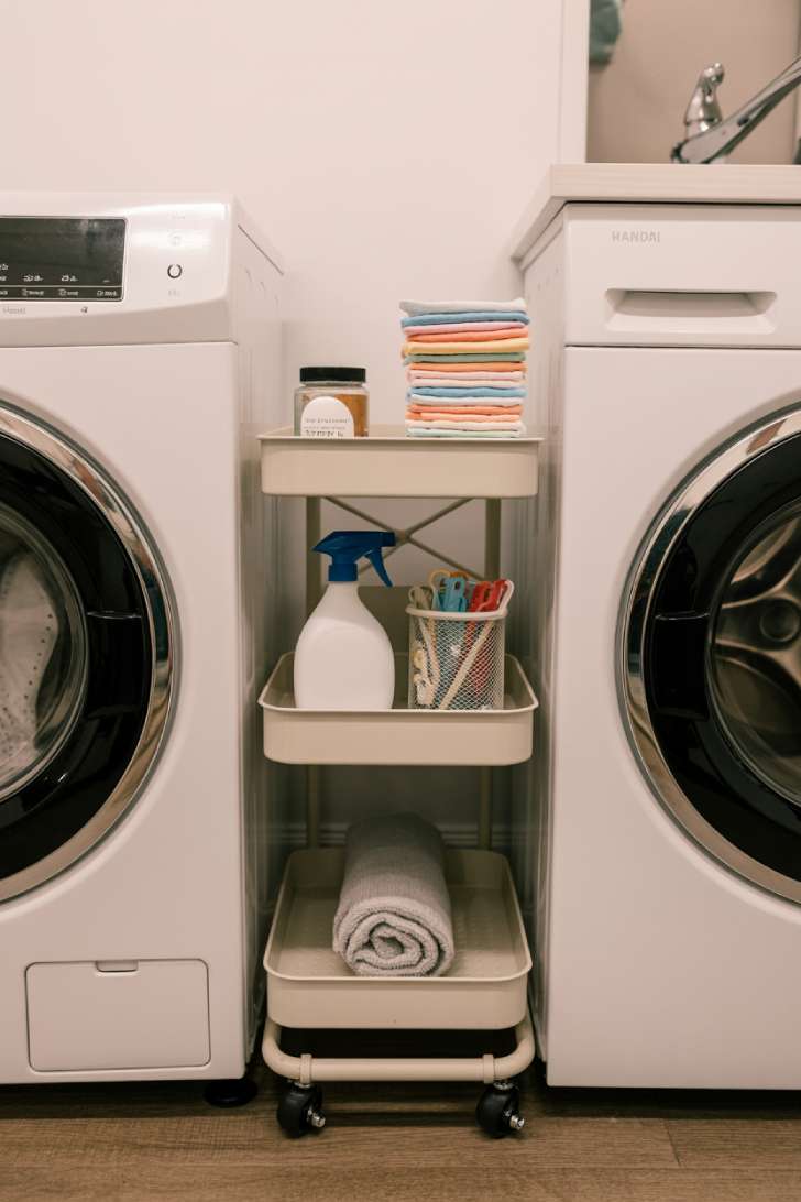 A slim white rolling cart with three shelves tucked between a washing machine and the wall, holding laundry supplies