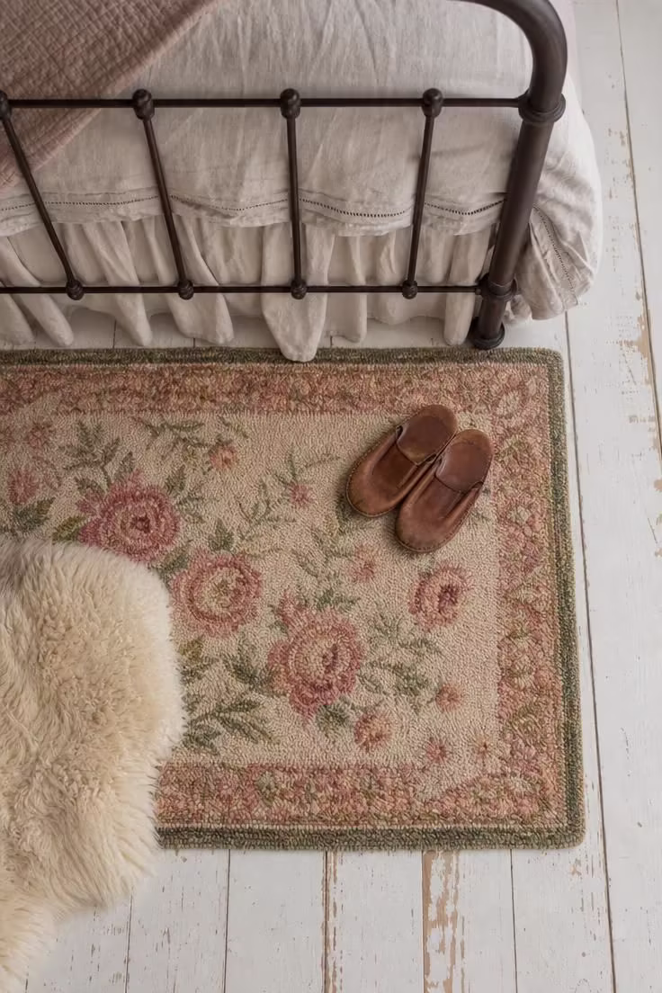 A faded floral hand-hooked wool rug in rose, green, and cream on painted white floorboards beside an iron bed frame.