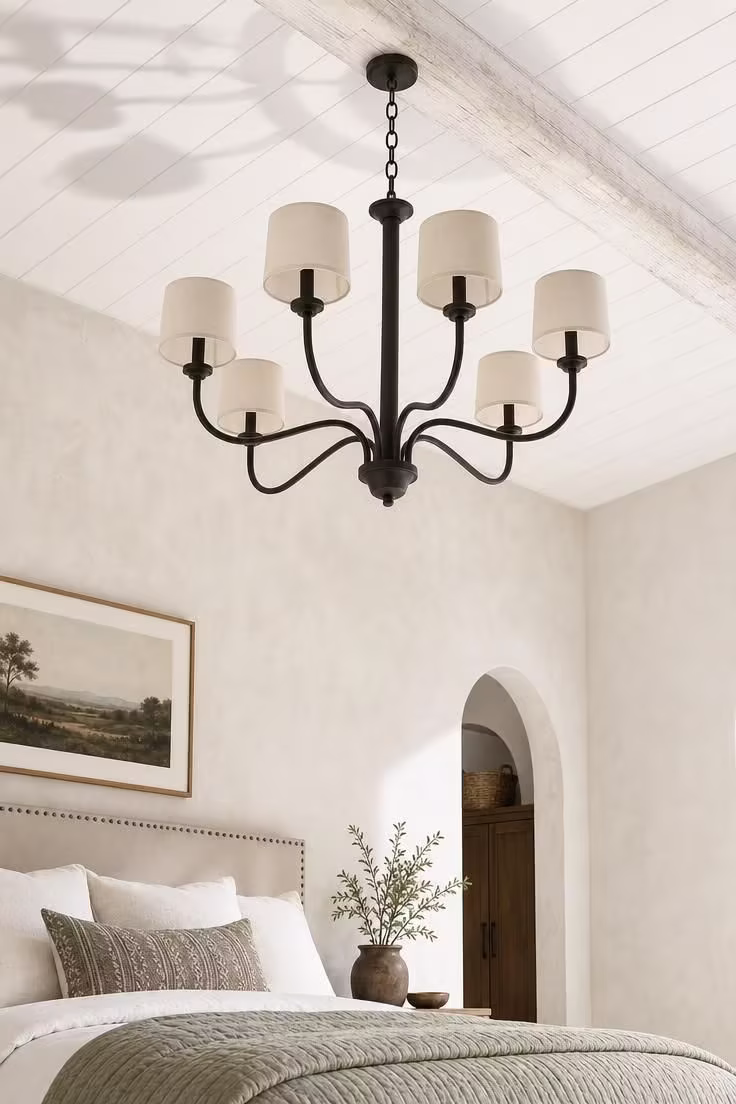 A six-arm wrought iron chandelier with cream linen drum shades hanging from a whitewashed beam against a beadboard ceiling.