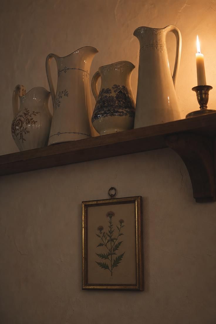 Four mismatched porcelain and ironstone pitchers displayed on a wood shelf rail with a single taper and a botanical print below.