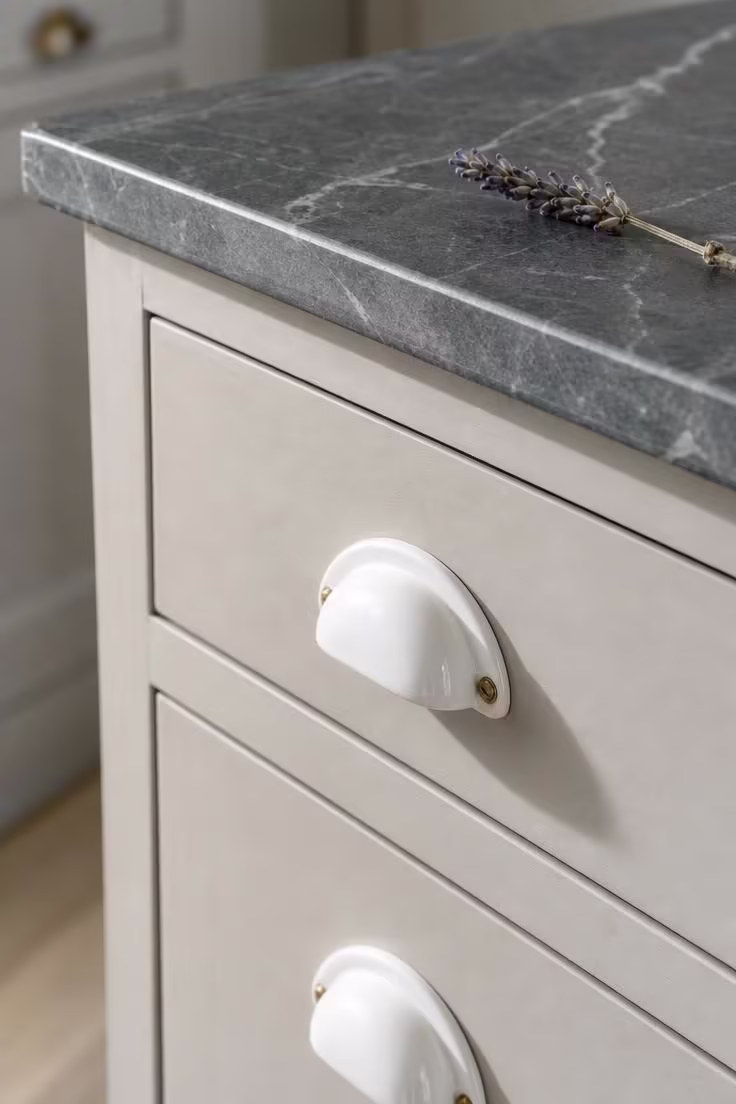 Porcelain bin pulls on putty-colored painted drawers with soapstone countertop in a vintage cottage kitchen