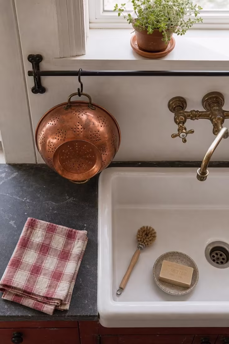 Copper colander hanging from a wrought iron S-hook above a white ceramic sink in a bright vintage cottage kitchen