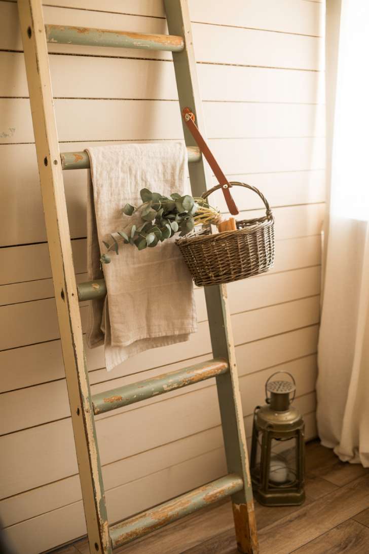 12 Timeless Vintage Laundry Room Decor Ideas to Steal Now 9 A weathered wooden ladder with old paint layers leaning against white shiplap in a vintage laundry room, styled with linen, a wicker basket, and dried eucalyptus