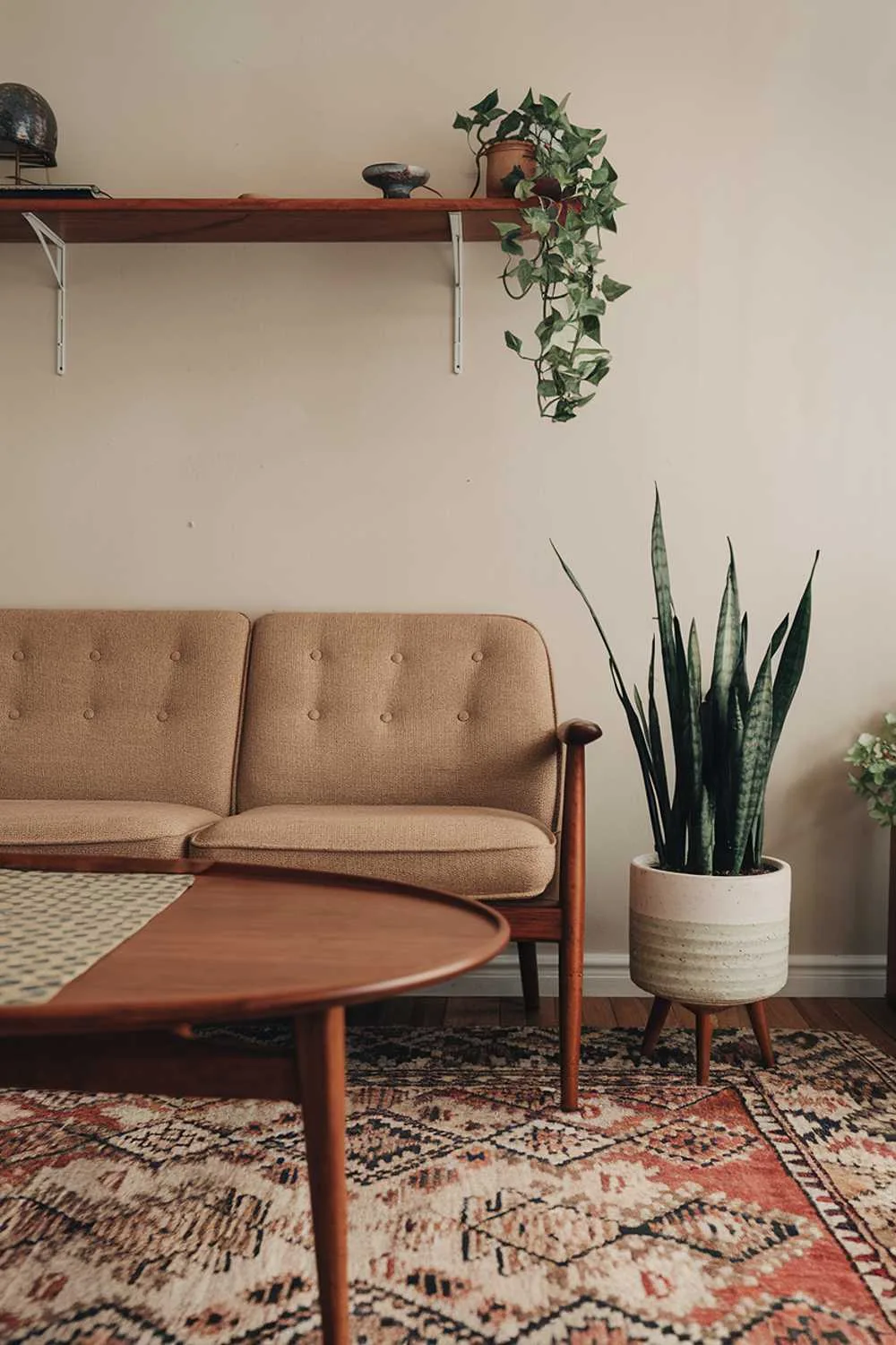 A vintage Scandinavian living room home decor. There's a beige sofa with wooden legs and a patterned rug beneath it. A wooden coffee table with a round top and a patterned surface is placed near the sofa. A green plant with a white pot sits on the floor near the coffee table. A wooden shelf with a few decorative items is mounted on the wall above the sofa. The wall is painted in a light, warm hue.