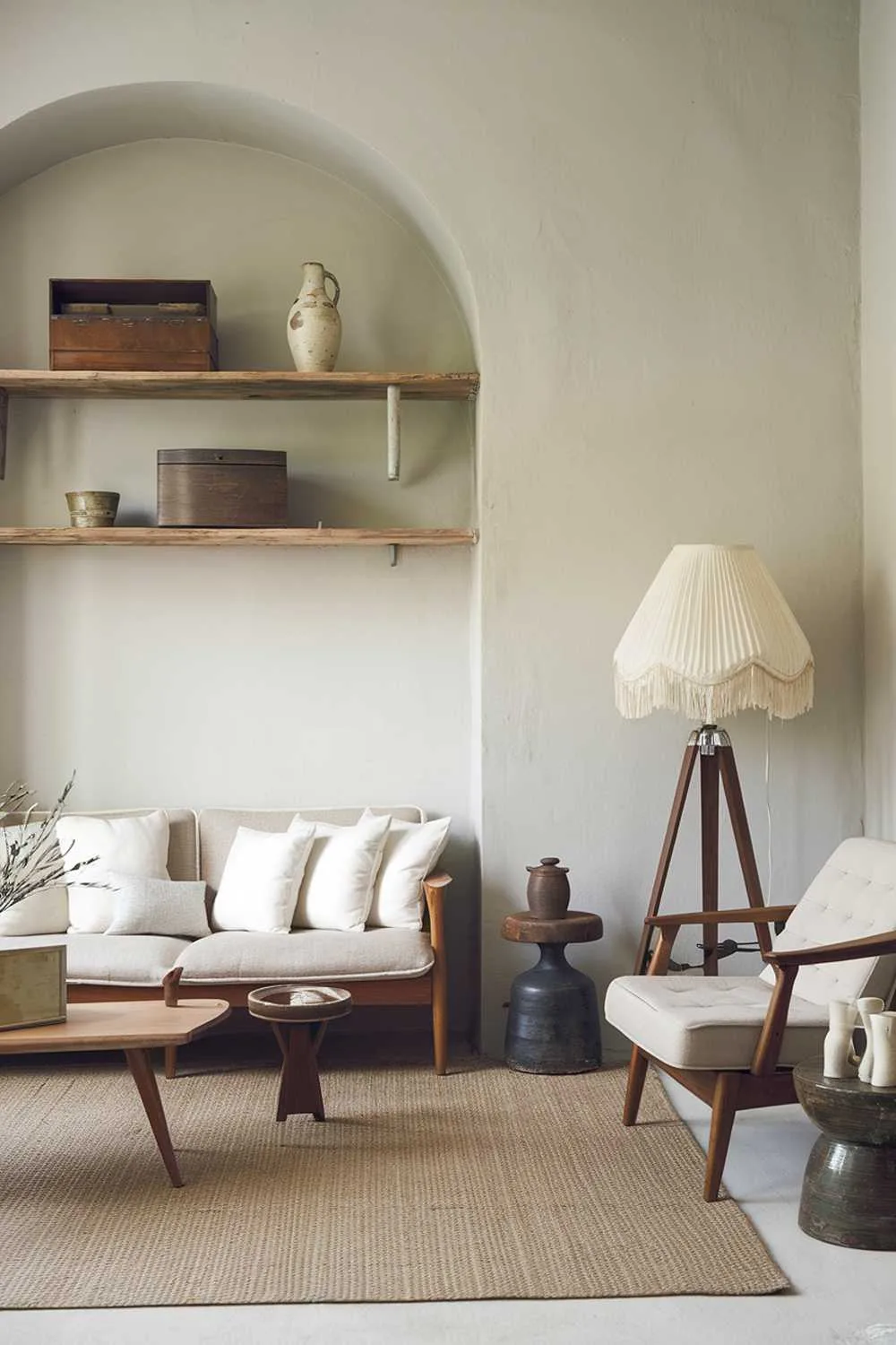 A Scandinavian living room with a vintage touch. The room has a beige sofa with white cushions and a few wooden furniture pieces. There's a vintage lamp with a white shade and a wooden stool. The walls are painted in a light gray color. The floor is covered with a beige rug. There are a few vintage items, such as a wooden box and a ceramic vase, placed on a wooden shelf above the sofa.