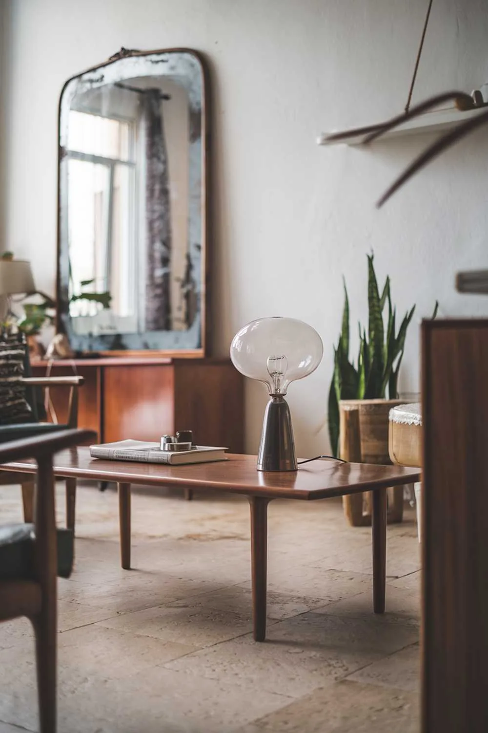 A Scandinavian-style living room with vintage elements. There is a wooden coffee table with a vintage lamp on top. The wall has a large vintage-style mirror. There's a wooden chair and a plant near the table. The floor is made of beige tiles. The room has soft lighting.