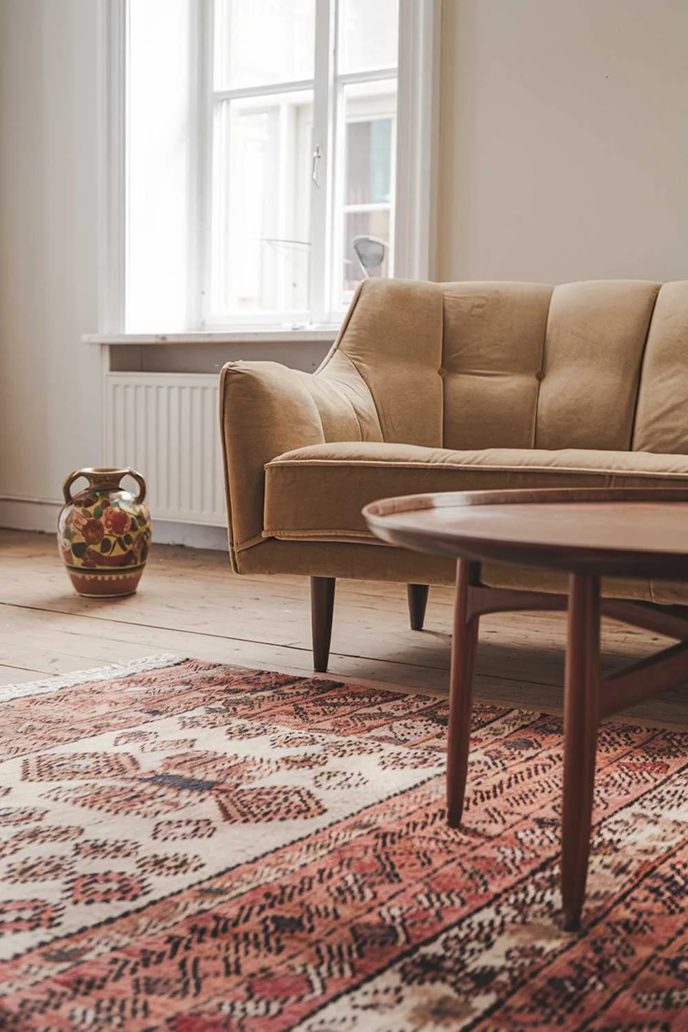 A Scandinavian living room with vintage finds. There's a patterned rug on the wooden floor. A beige velvet sofa with a mid-century modern design is placed near the window. A wooden coffee table with a round top and four legs is placed in front of the sofa. There's a vintage ceramic vase with a floral pattern on the wooden floor near the sofa. The walls are painted in a light color.