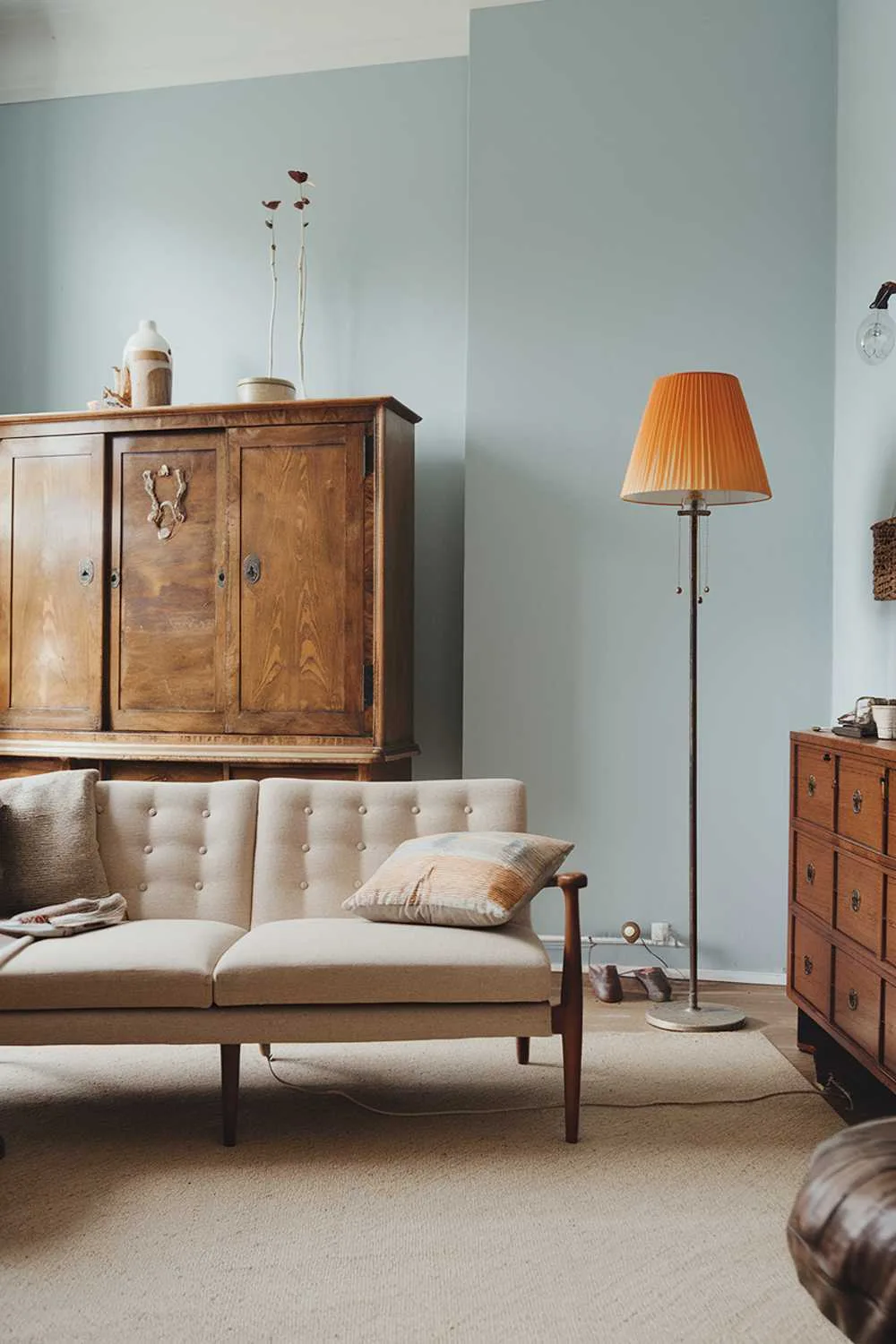 A Scandinavian living room with vintage finds. The room has a beige sofa with wooden legs and a few throw pillows. There's a vintage lamp with a orange shade near the sofa. On the opposite wall, there's a vintage wooden cabinet with doors. The floor is covered with a beige rug. The walls are painted in a light blue hue.