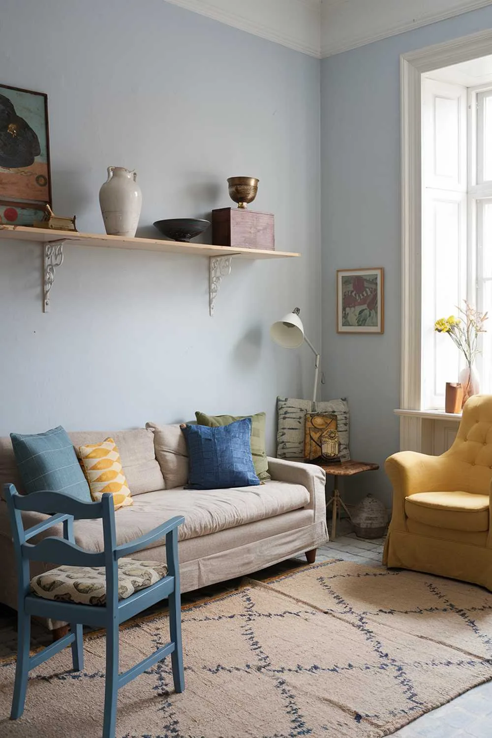 A Scandinavian living room with vintage finds. There is a beige sofa with cushions in blue, green, and yellow. A yellow armchair is placed near the window. There is a blue chair with a patterned cushion. A white shelf is placed above the sofa, holding a white vase and a few vintage items, such as a lamp, a bowl, and a wooden box. The floor is covered with a beige rug with a pattern. The walls are painted in light blue.