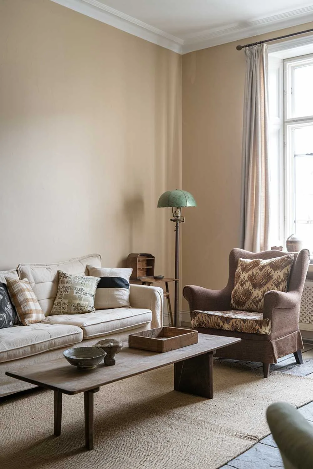 A Scandinavian living room with a vintage touch. There is a beige sofa with cushions in various patterns. There is a brown armchair with a patterned cushion. A wooden coffee table sits between the sofa and the armchair. The walls are painted beige. There are a few vintage items, such as a wooden box and a ceramic bowl, placed on the coffee table. A vintage lamp with a green shade stands near the window. The floor is covered with a beige rug. The window has a curtain.