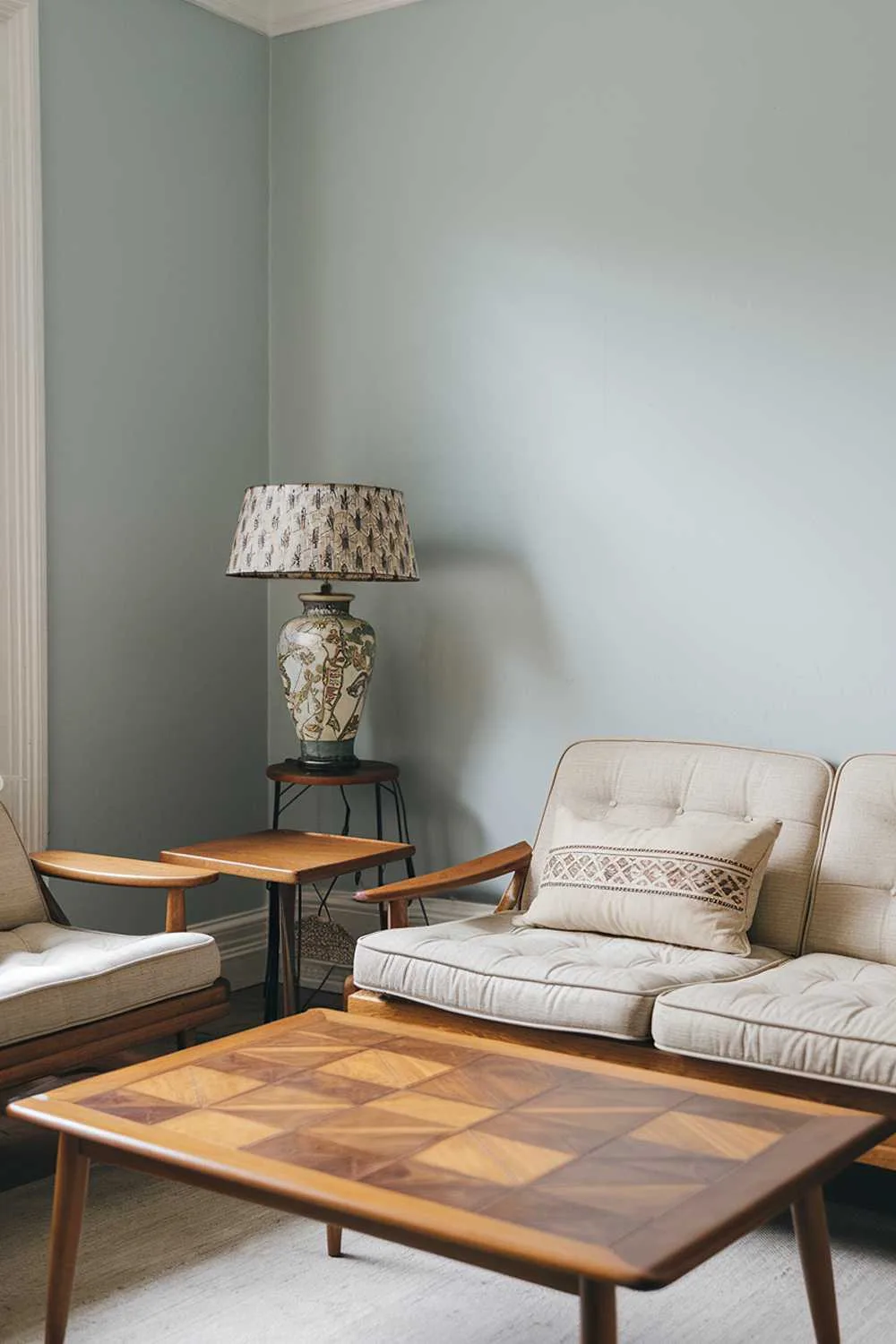 A Scandinavian living room with vintage furniture. There is a wooden coffee table with a geometric pattern and a beige couch with a few pillows. The walls are painted in a light blue color. There is a vintage lamp with a patterned shade and a wooden side table next to the couch.