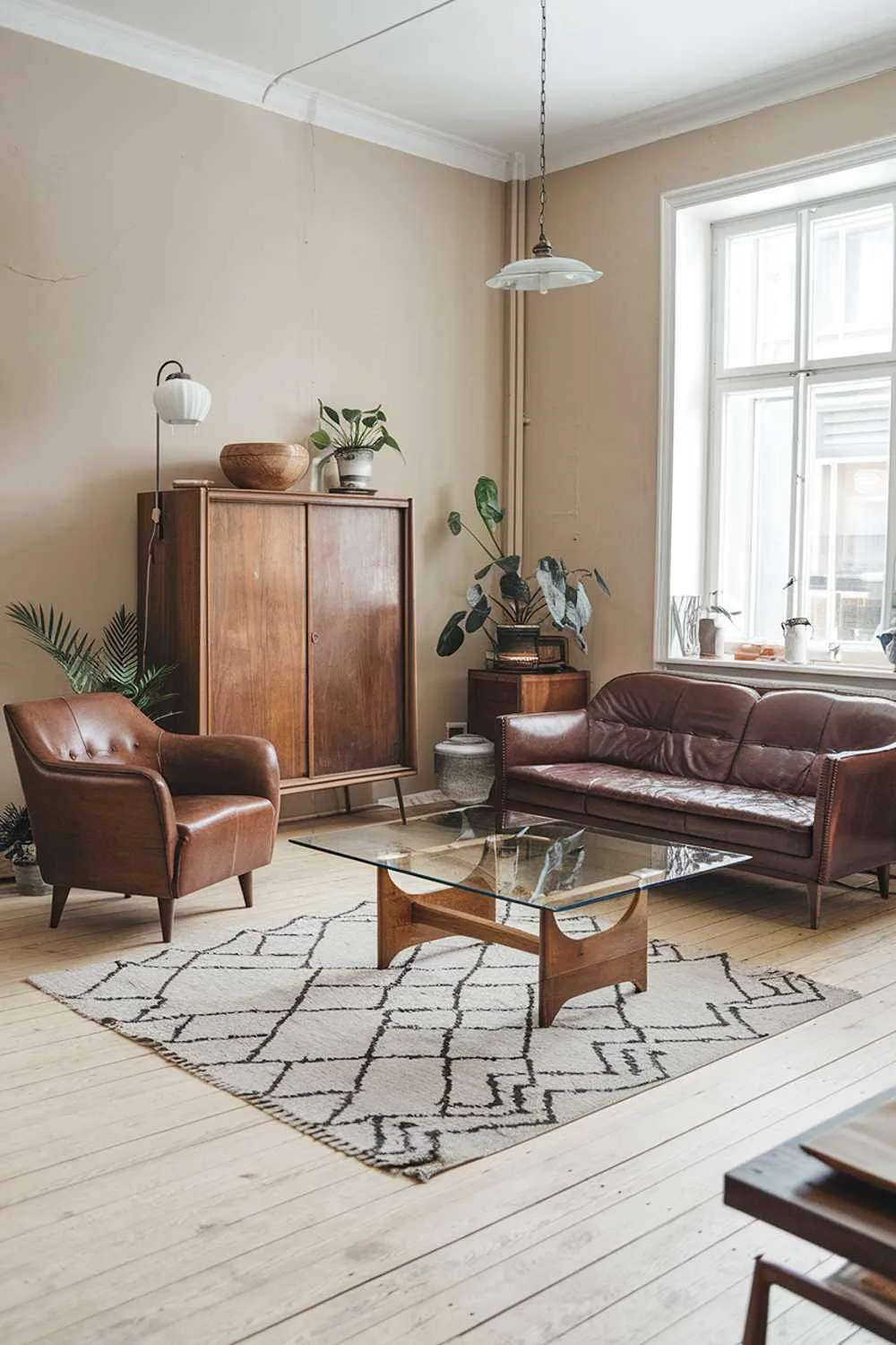 A Scandinavian living room with vintage furniture. The room has a light wooden floor and beige walls. There's a brown leather sofa and a matching armchair near the window. A glass coffee table with a wooden base sits in front of the sofa. A vintage wooden cabinet is against the wall. A rug with a geometric pattern is in front of the cabinet. A potted plant is near the cabinet. The room has a vintage lamp and a few decorative items, including a wooden bowl and a ceramic pot.