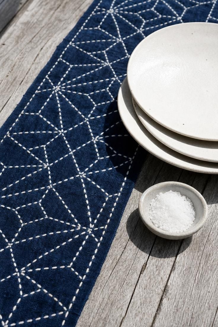 13 Earthy Wabi Sabi Dining Area Ideas That Feel Like Home 13 A hand-stitched indigo sashiko table runner on a weathered silver-gray teak dining table with white stoneware plates in bright midday light.