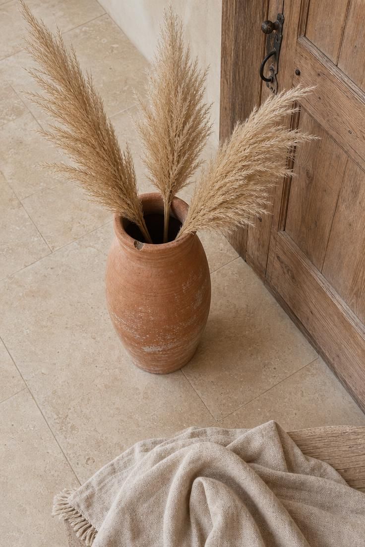 Chipped terracotta floor vase with dried pampas grass plumes beside a reclaimed wood front door in a wabi sabi entryway.