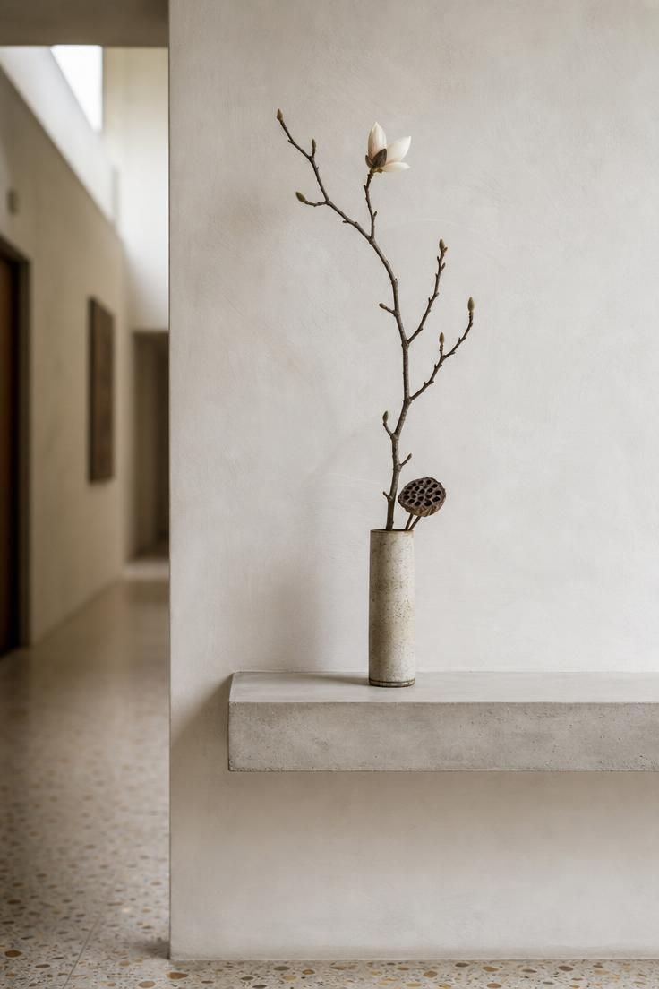 Single ikebana arrangement in a stoneware cylinder on a raw concrete ledge in a wabi sabi entryway.