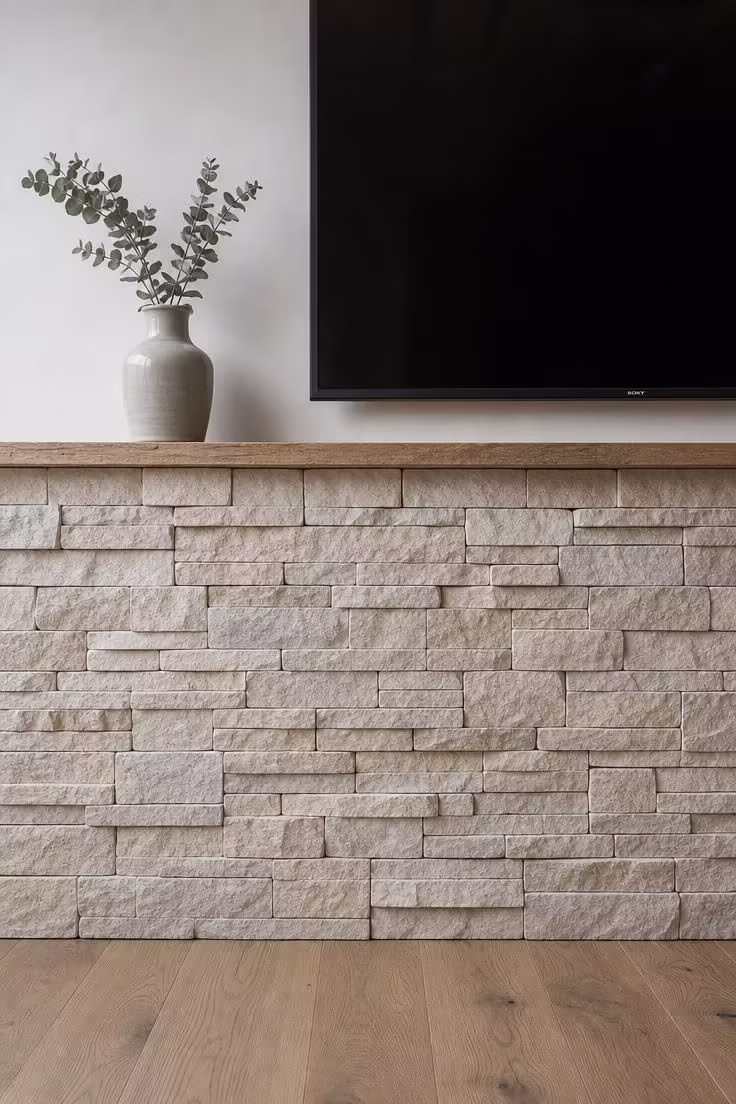 Close-up of stacked limestone ledger panels on the lower half of a TV wall with a raw-edge wood shelf, clay vessel, and dried eucalyptus in cool overcast light.