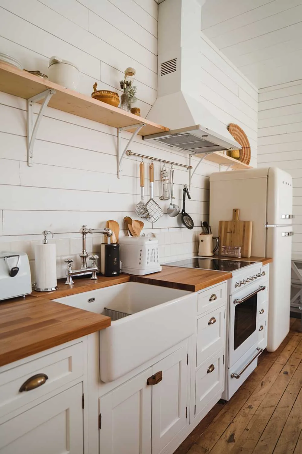 A white and wood kitchen decor with a farmhouse sink, a range hood, a white refrigerator, and wooden counters. There are also a few kitchen appliances and utensils placed on the counter. The floor is made of wood.