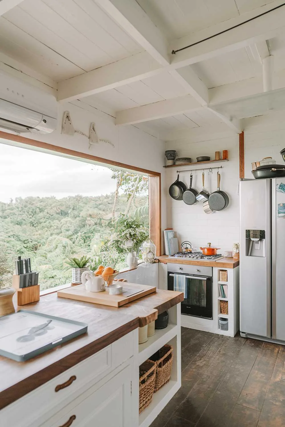 A kitchen with a white and wood decor. The kitchen has a modern design with a large window overlooking a lush green landscape. The countertop is made of wood and has a few items on it. There's a white fridge with a water dispenser. The floor is made of wood. A few pots and pans are hanging above the stove.