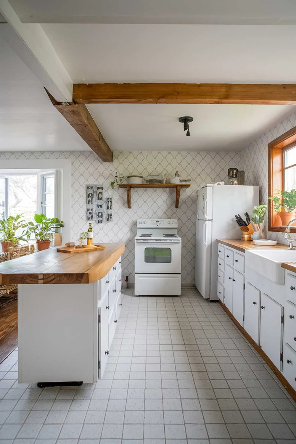 A white and natural wood kitchen decor. The kitchen has a white island with a natural wood counter and white cabinets. There is a white stove, a white fridge, and a white sink. The natural wood elements include the island counter, a wooden shelf above the fridge, and a wooden beam in the ceiling. The floor is tiled with white and gray squares. There are potted plants near the window. The walls have white and gray patterned wallpaper.