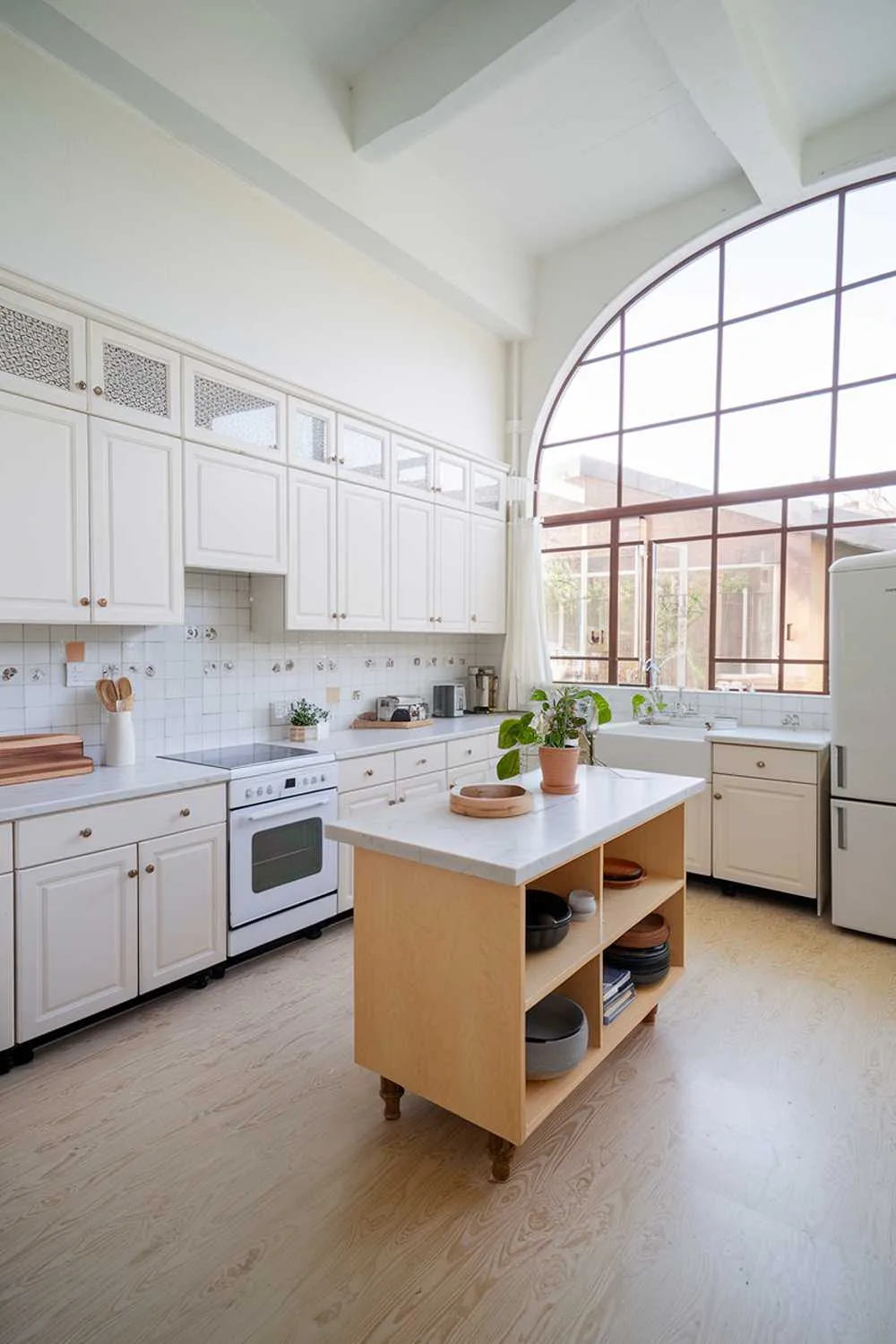 A kitchen with white cabinets and a light wood floor. There's a large window near the ceiling, allowing for natural light. The countertops are made of white marble. There's a white refrigerator, a white oven, and a white sink. There's a light wood island in the middle of the room, with a white countertop. The island has a few items, including a wooden bowl and a potted plant. The backsplash has a white wall and a few decorative tiles. There's a white chair near the window. The floor has a light wood pattern.