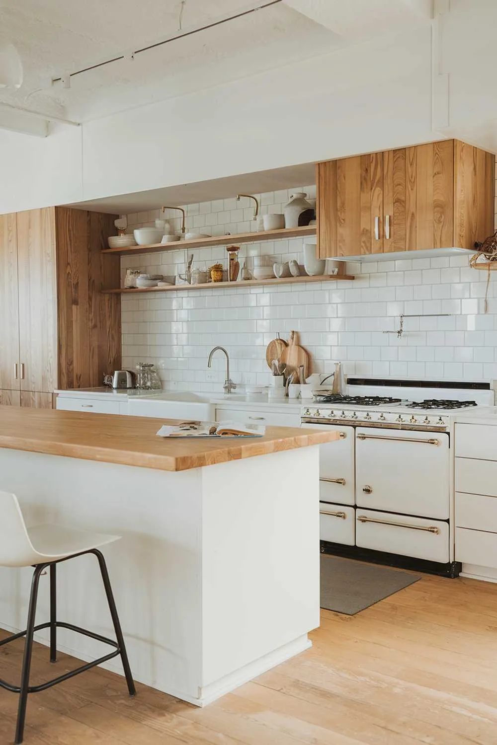 A white and light wood kitchen decor. The kitchen has a white island with a wooden countertop. There is a white stove with wooden cabinets above it. The floor is made of light wood. There is a white chair near the island.