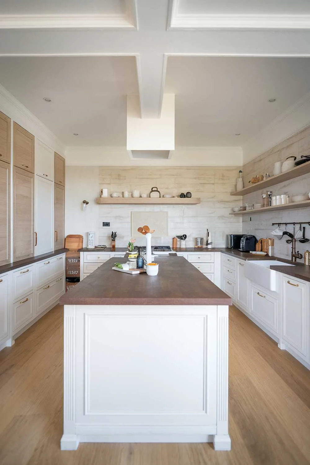 A kitchen with a white and light wood decor. The kitchen has a white island in the center with a dark brown countertop. There are white cabinets along the walls, with some of them having light wood doors. The backsplash has a light wood pattern. There is a white sink with a dark brown faucet. The floor is made of light wood. There are various kitchen appliances, pots, and utensils in the kitchen.