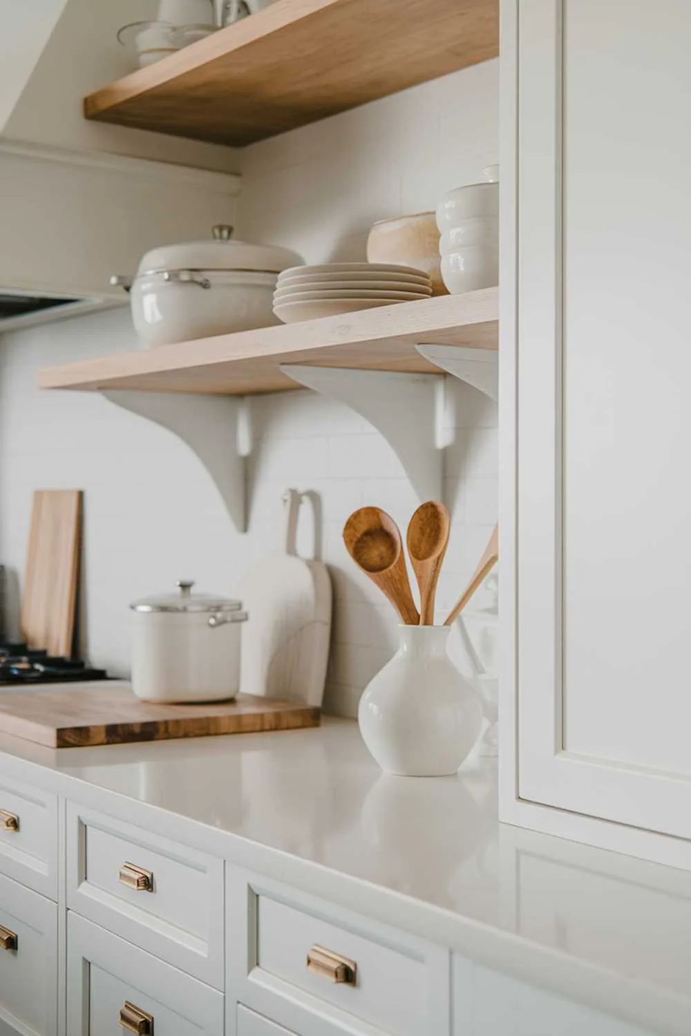 A white and light wood kitchen decor. The background reveals a white cabinet with light wood shelves and a white countertop. There are white pots and a wooden spoon on the counter. The cabinet has a few white items and a light wood item. The lighting is soft.