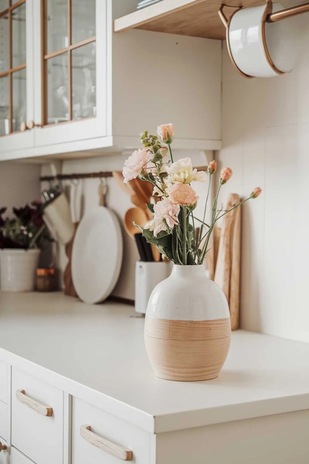 A white and light wood kitchen decor. There is a white vase with light wood details on a white counter. The counter has a few fresh flowers and a few utensils. The background contains a white cabinet with light wood details and a few hanging pots. The overall image has a warm and cozy ambiance.