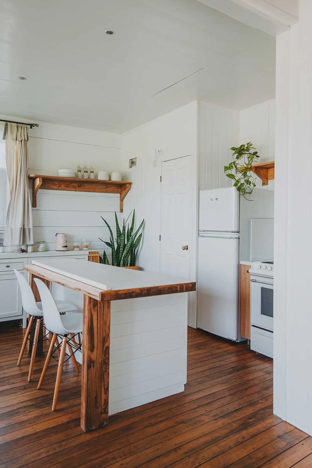 A white kitchen with wood accents. There's a wooden floor, a wooden island with a white counter, and a wooden shelf above the door. There's a white cabinet, a white fridge, and a white oven. There are white chairs at the island. There's a potted plant near the fridge. The walls are white. The room has a window with curtains.