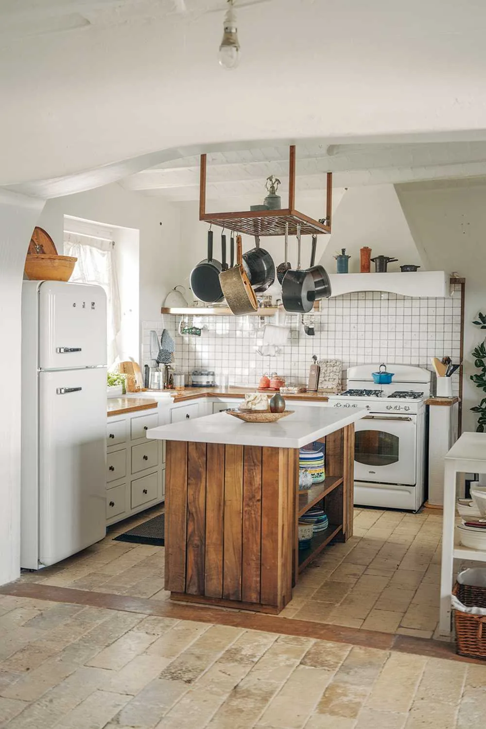 A white kitchen with a wood island. The kitchen has a white cabinets, a white refrigerator, and a white oven. There is a wooden island in the middle of the kitchen, with a white counter on top of it. The floor is made of beige tiles. There are pots and pans hanging above the island. The lighting is bright.