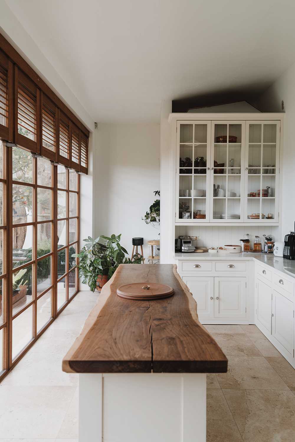 A white kitchen with a wood island. The kitchen has a large window with wooden blinds on the left, allowing natural light to fill the space. There is a white cabinet with glass doors on the right, housing various cooking utensils. The wood island has a circular wooden object on it. There's a potted plant near the window. The floor is made of beige tiles.