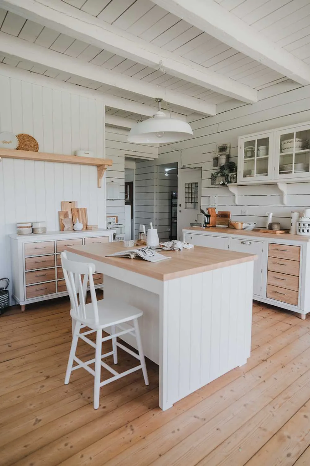 A white and light wood kitchen with a rustic decor. The kitchen has a white island with a light wood countertop, a white cabinet, and a light wood wall. There's a white chair near the island. On the opposite side of the kitchen, there's a white cabinet with light wood drawers. There's a white wall with a light wood shelf and a white light fixture hanging above the island. The floor is made of light wood planks.