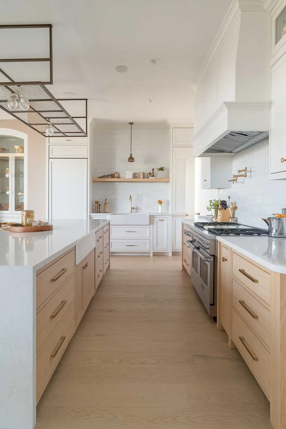 A modern kitchen with a white and light wood decor. The kitchen has a large island with a white quartz countertop and light wood cabinets. There is a white sink with a gold faucet in the background. There are white cabinets and a light wood floor throughout the kitchen. There is a pot rack above the island. The lighting is bright.