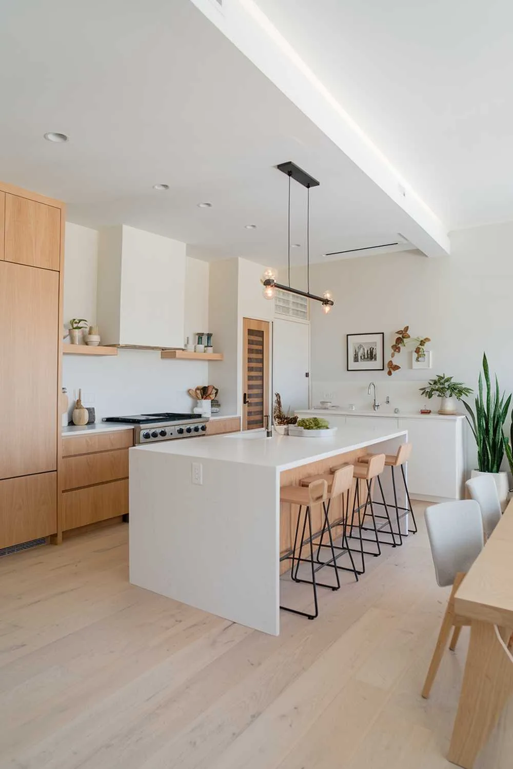 A modern kitchen with a white and light wood decor. The kitchen has a white island with a few stools and a white cabinet. The walls are painted white and have a few floating shelves. There's a light wood floor and a light wood cabinet on the left side. The kitchen has a few potted plants. The lighting is bright.