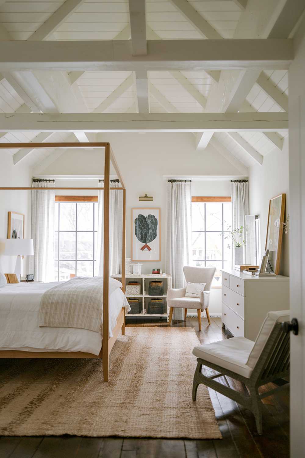  A cozy japandi-style bedroom with a white color scheme. The room features a high ceiling, wooden beams, and a wooden floor. There's a white bed with a wooden frame, a white dresser, and a white chair. A beige rug is placed under the bed. The walls are adorned with a few pieces of artwork.