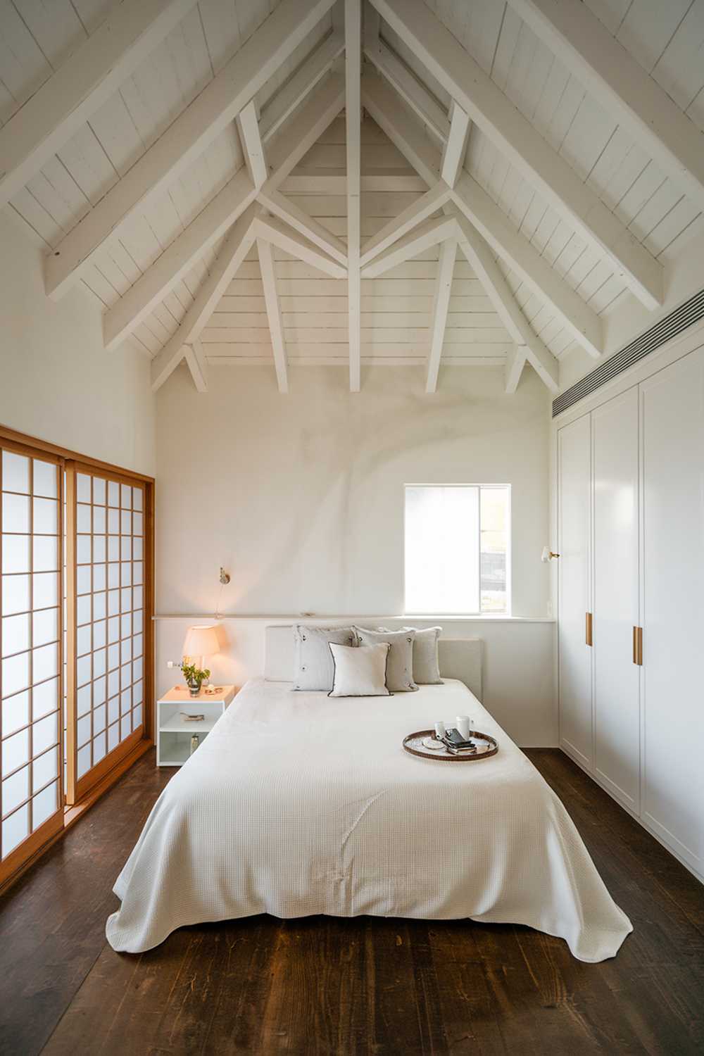 A modern white japandi bedroom design. The room has a high ceiling with wooden beams. There is a king-size bed with a white cover and a few pillows. Next to the bed, there is a small white nightstand with a lamp. On the opposite wall, there is a white wardrobe. The floor is made of dark wood. There is a large window with wooden frames, allowing natural light to fill the room.